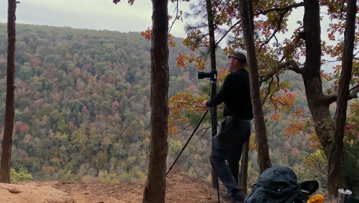 Photographer on tripod shooting landscape from forest overlook in autumn.