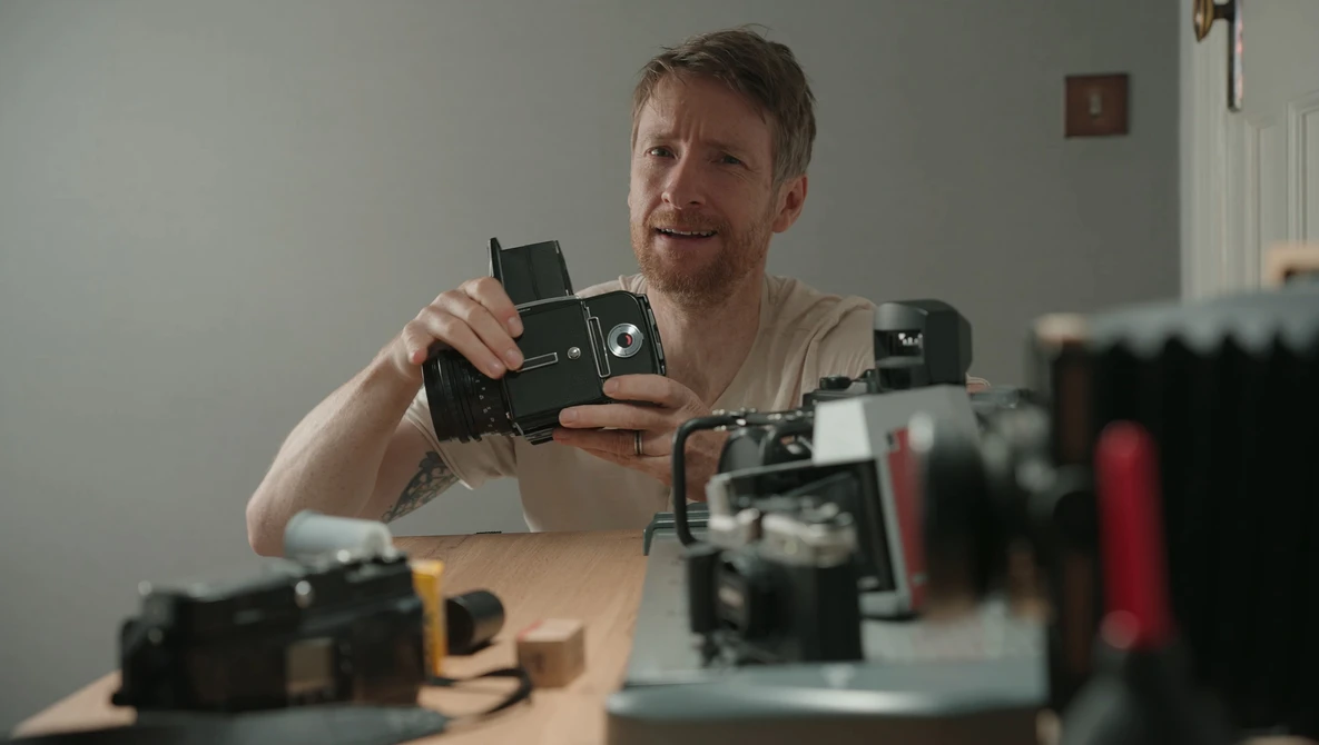 Photographer examining a vintage film camera while surrounded by other classic cameras on a wooden desk.
