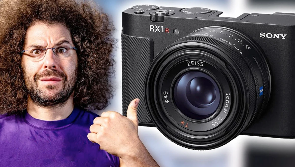 Man in purple shirt with curly hair and glasses gesturing toward a Sony RX1 II compact camera.