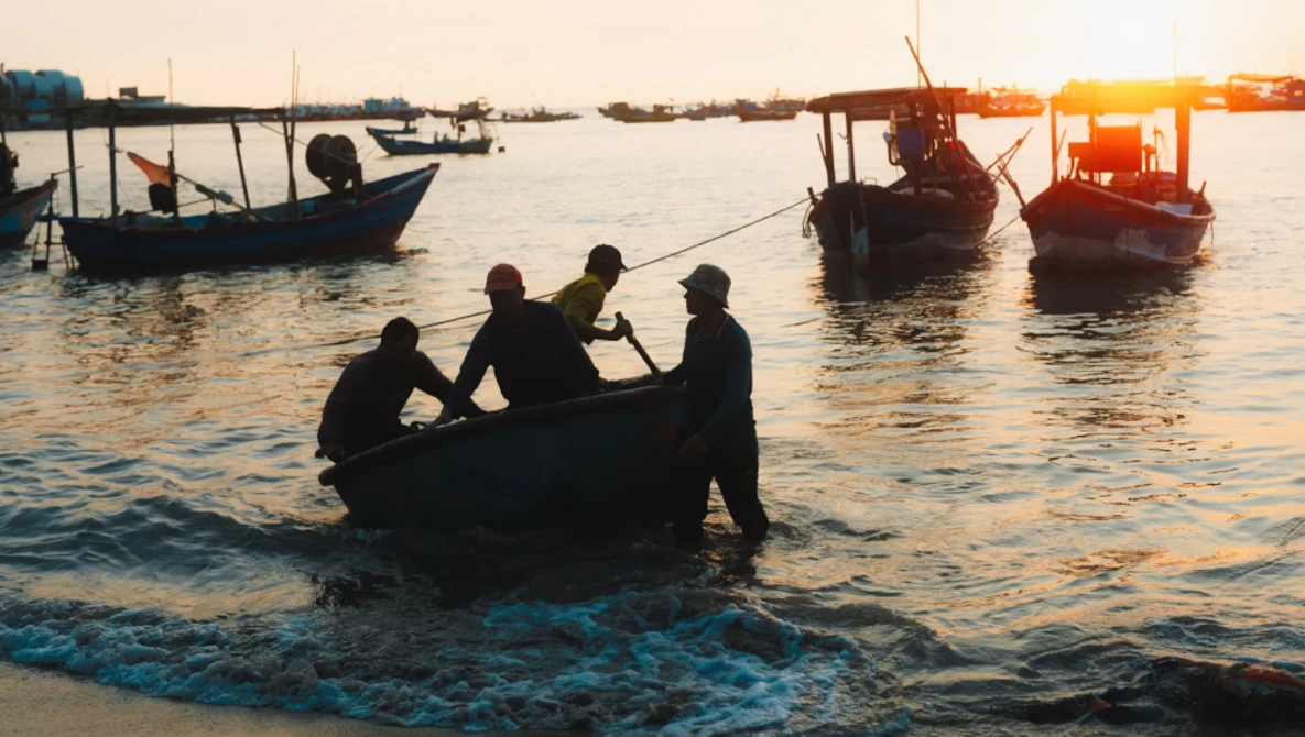 Silhouetted fishermen wade in shallow water carrying a net at golden hour with fishing boats anchored in the background.