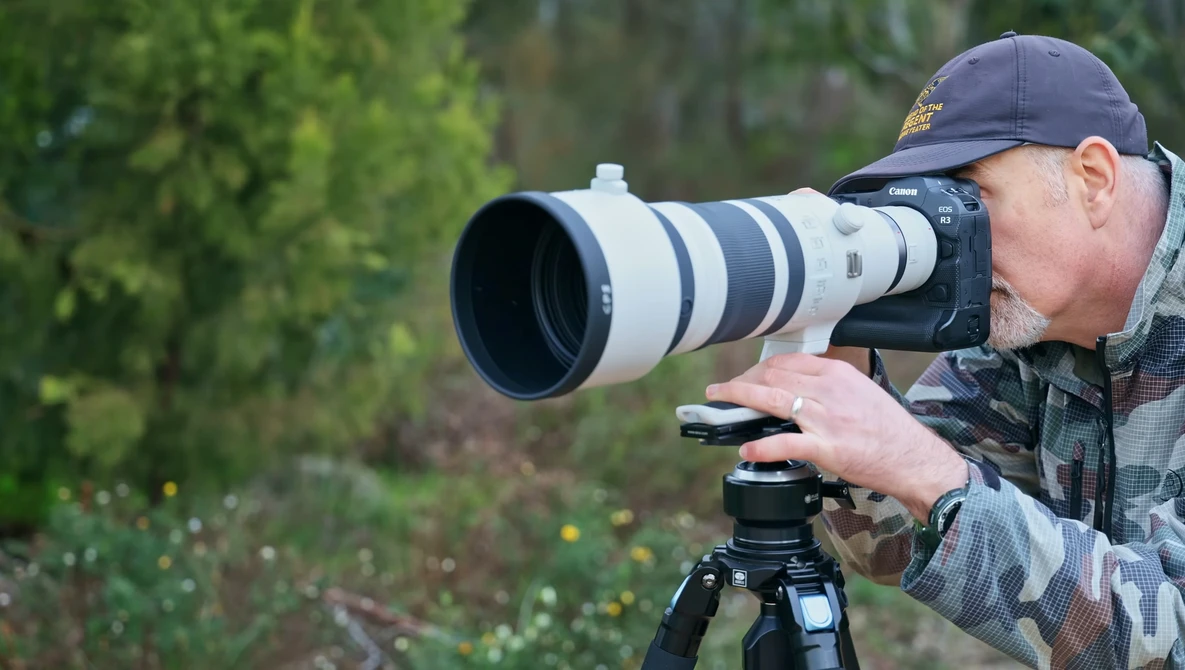 Photographer holding a telephoto lens on a tripod-mounted camera, focused on distant subject in forested setting.