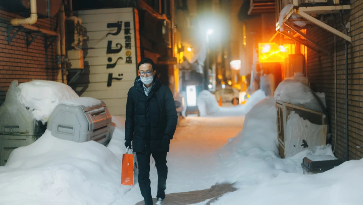 Person standing in snowy alley at night holding red shopping bag, wearing mask and dark coat.