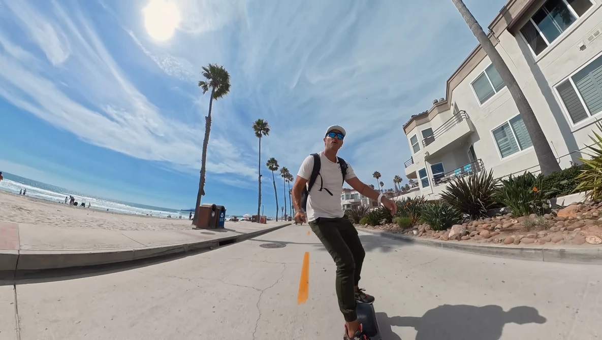 Person riding an electric skateboard on a coastal path with palm trees and beach buildings visible.