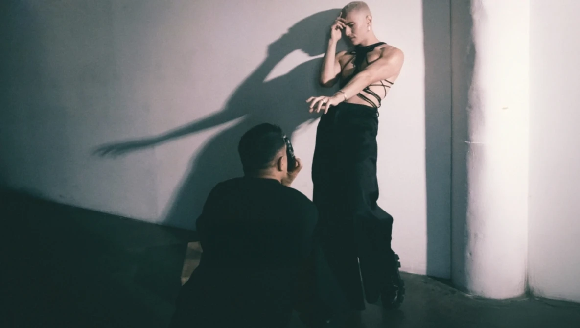 Shirtless man against white wall with dramatic shadow of another person cast across his torso.