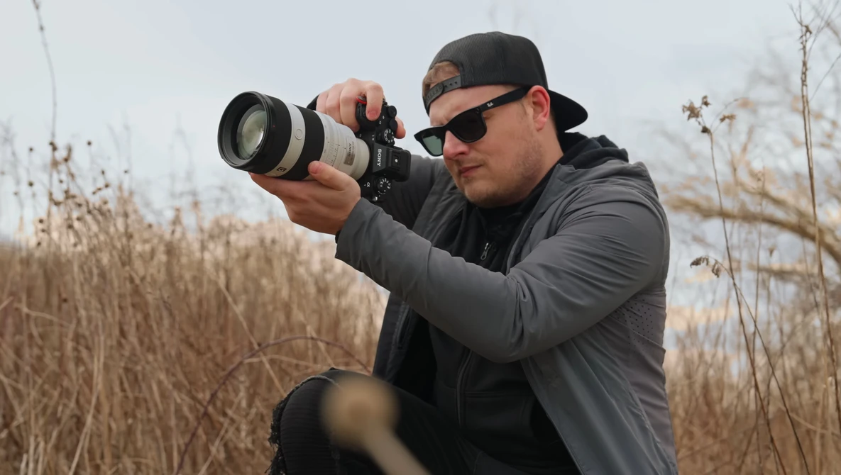 Photographer holding a telephoto lens camera while looking through the viewfinder in an open field.