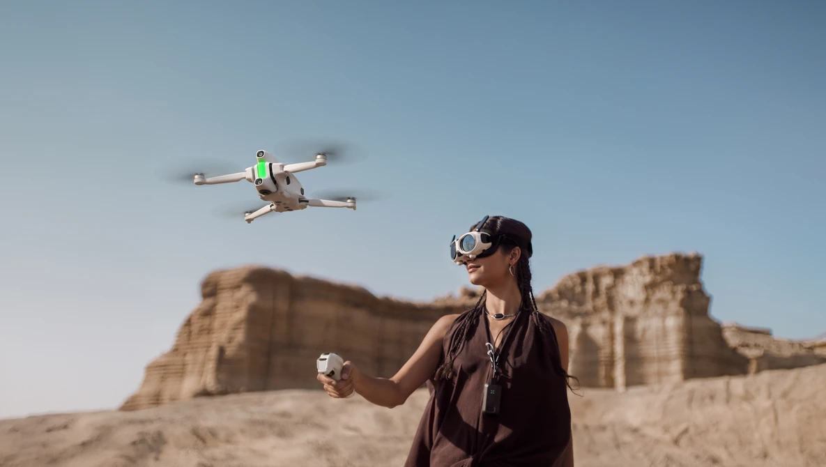 Operator wearing VR headset controls a small quadcopter drone in flight above desert terrain.