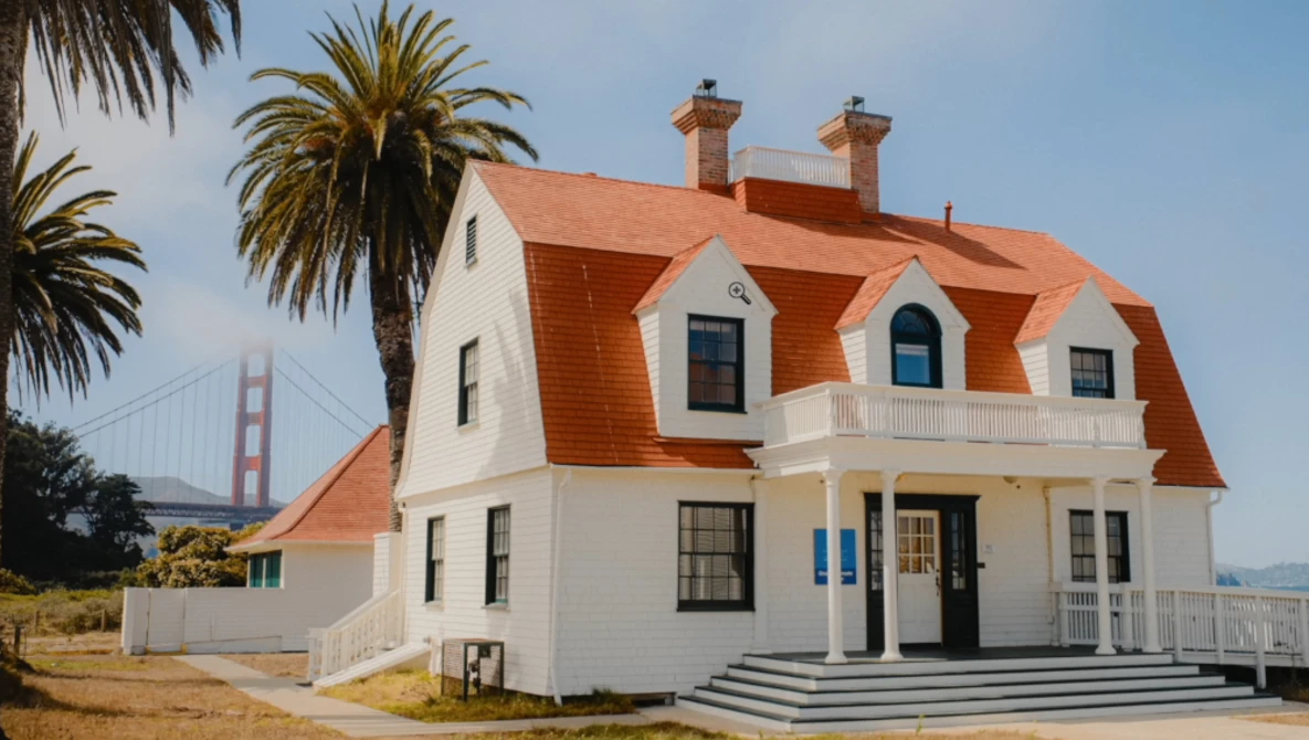 White Victorian-style house with distinctive orange-red mansard roof and dormer windows, palm trees nearby, suspension bridge visible in background.