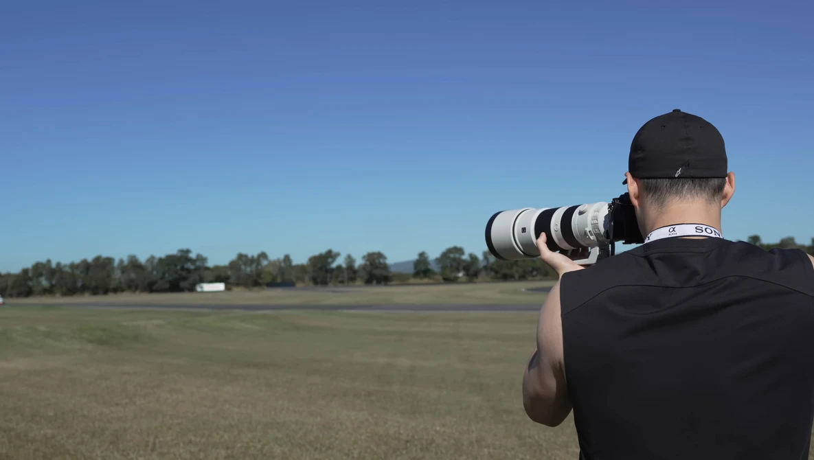 Photographer from behind holding a telephoto lens, shooting across an open field under clear blue sky.
