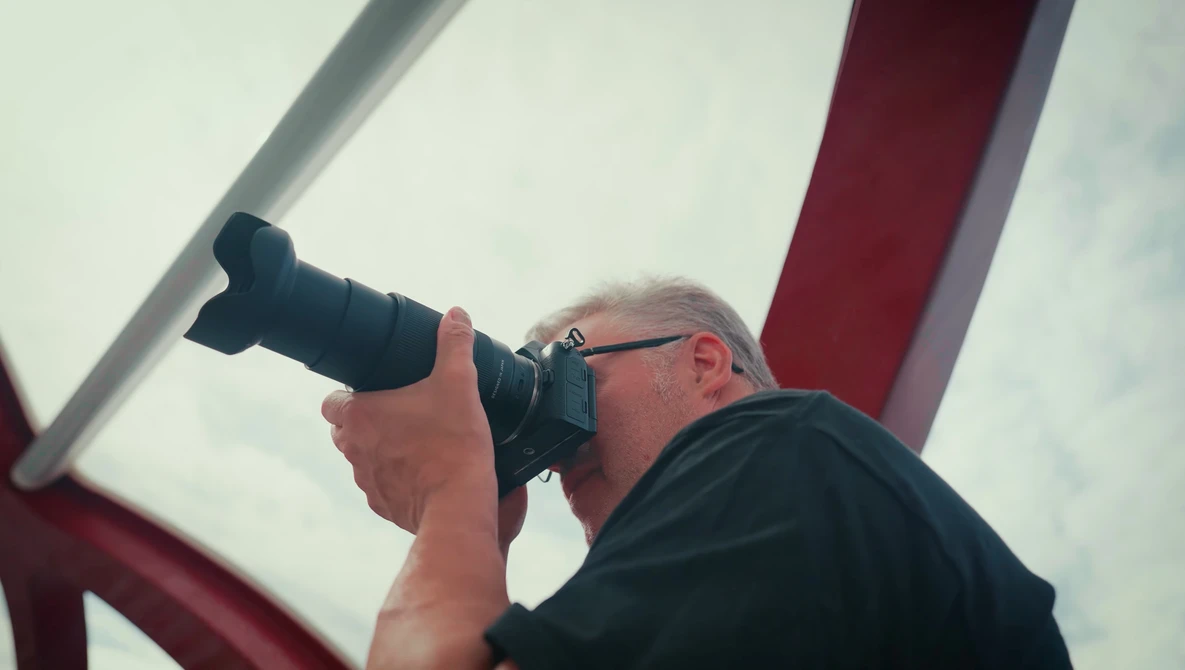 Photographer holding telephoto lens up to eye while shooting outdoors against cloudy sky.