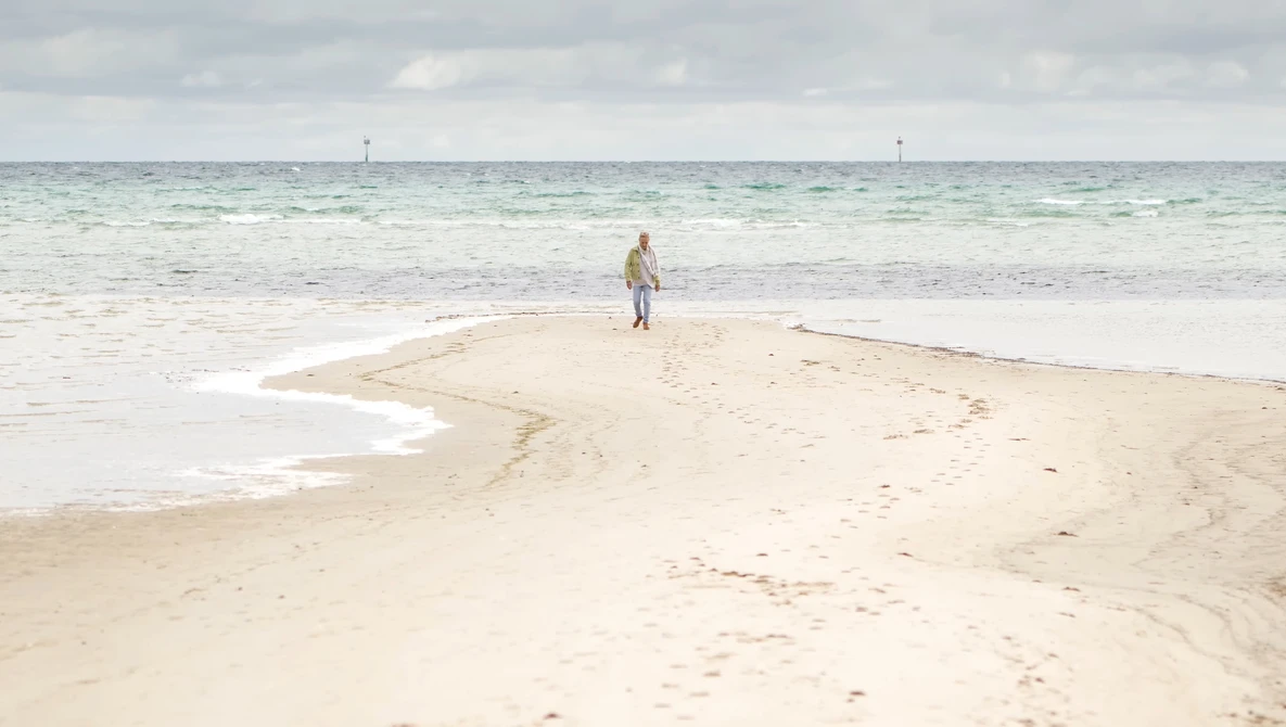 Lone figure standing on a curved sandbar extending into shallow turquoise water.
