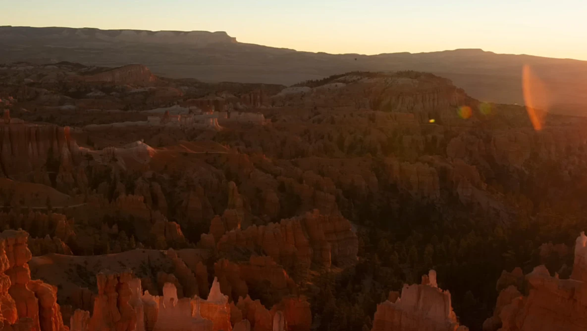 Golden sunrise light illuminates layered hoodoos and forested ridges in a vast canyon landscape.