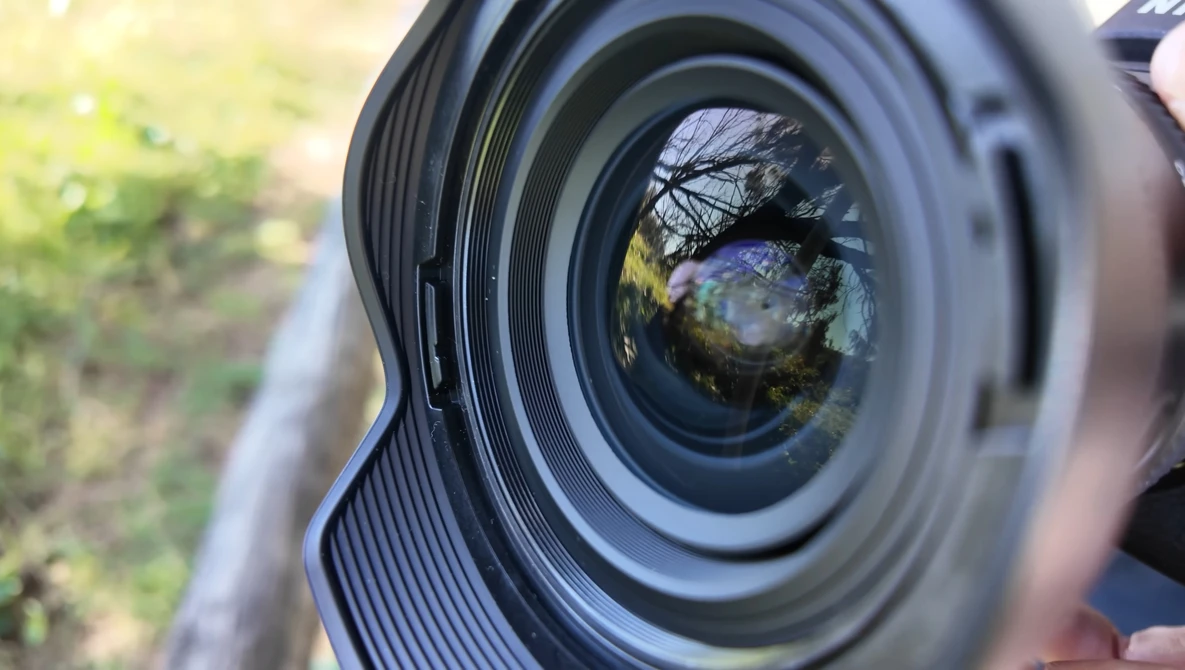 Close-up of a camera lens front element with shallow depth of field, showing tree branches and landscape reflected in the glass.