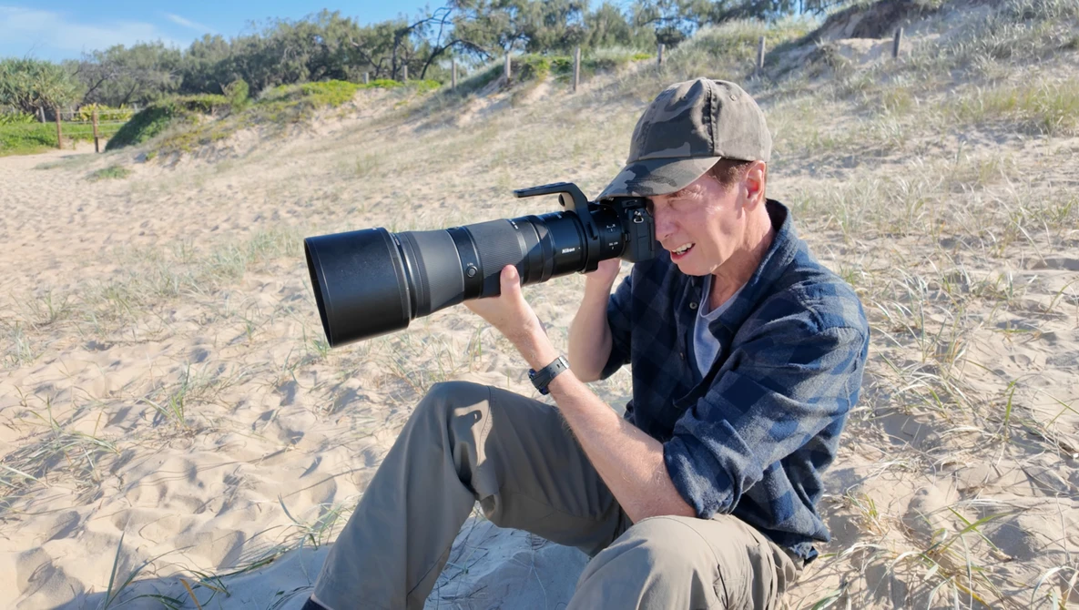Photographer seated on sand using a telephoto lens while shooting at a beach location.