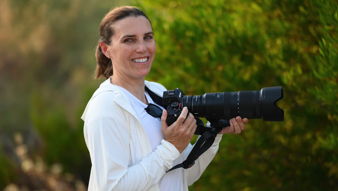 Photographer holding a professional camera with telephoto lens, smiling at camera against blurred green foliage background.
