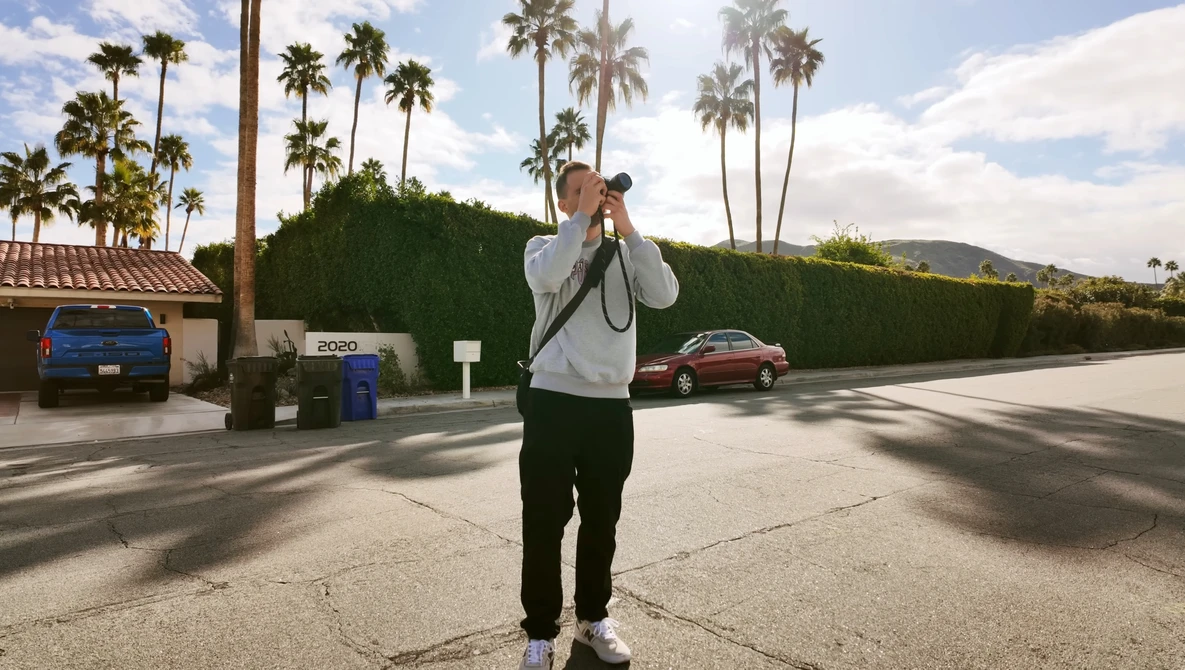 Photographer holding camera up to face while standing in a residential driveway with palm trees and mountains in background.
