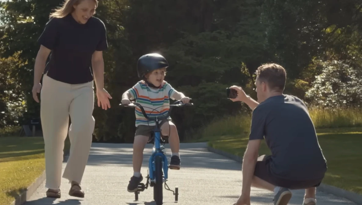 Woman supervises young child riding blue bicycle with training wheels on a paved path as man crouches nearby.
