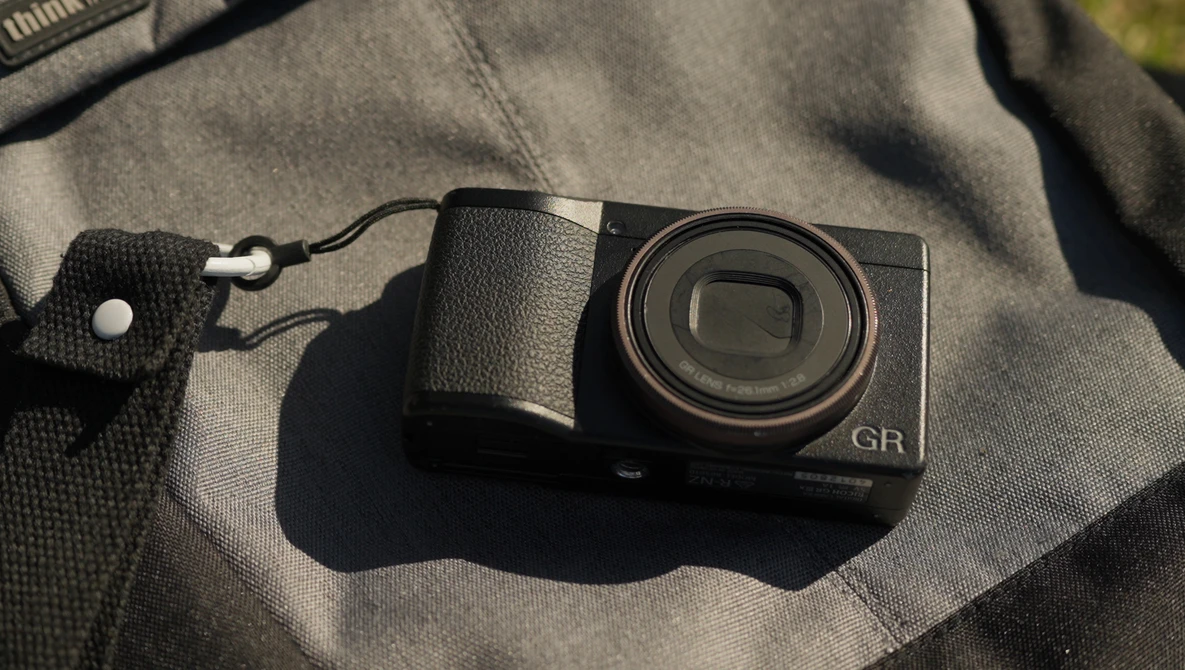 Ricoh GR compact digital camera with textured grip sitting on a gray fabric surface with a wrist strap attached.