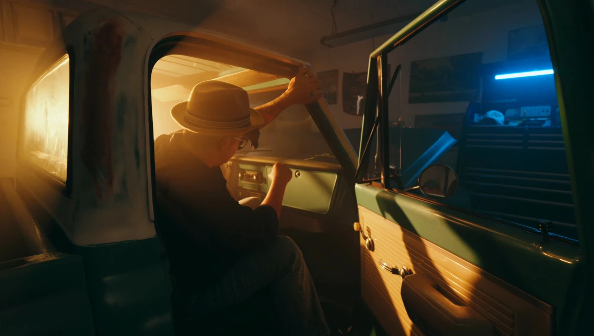 Silhouetted figure wearing a hat inside a vehicle at night, illuminated by warm and cool ambient light.