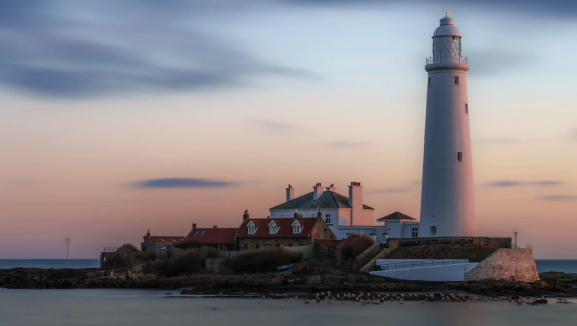 Lighthouse with attached keeper's house on rocky outcrop at twilight, warm sky with cool water below.