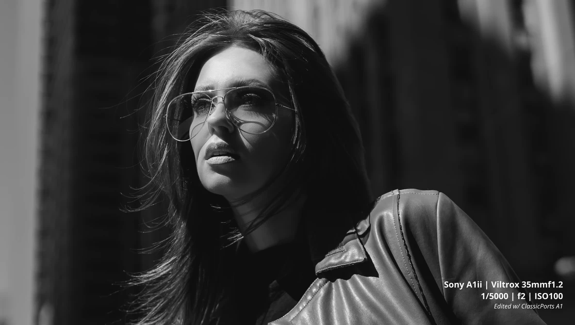 Person wearing clear-framed glasses and light-colored jacket, photographed in black and white with directional side lighting.