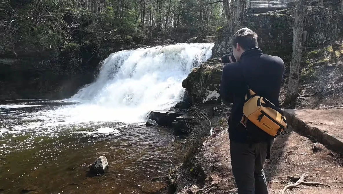 Photographer with camera and yellow bag documents a cascading waterfall in a forested stream setting.