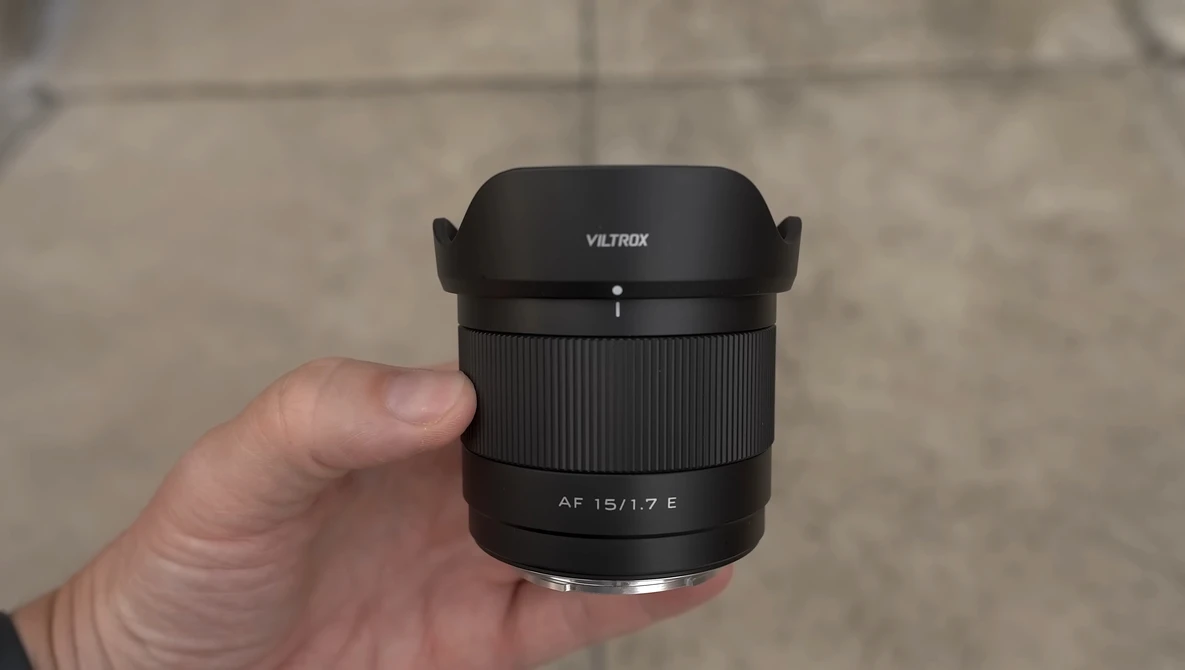Hand holding a Viltrox AF 15/1.7 E lens with hood attached against a concrete background.
