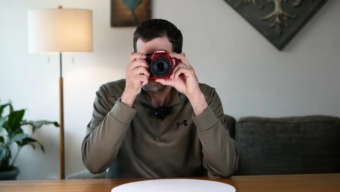 Photographer holding a red digital camera up to his face while seated at a wooden table.