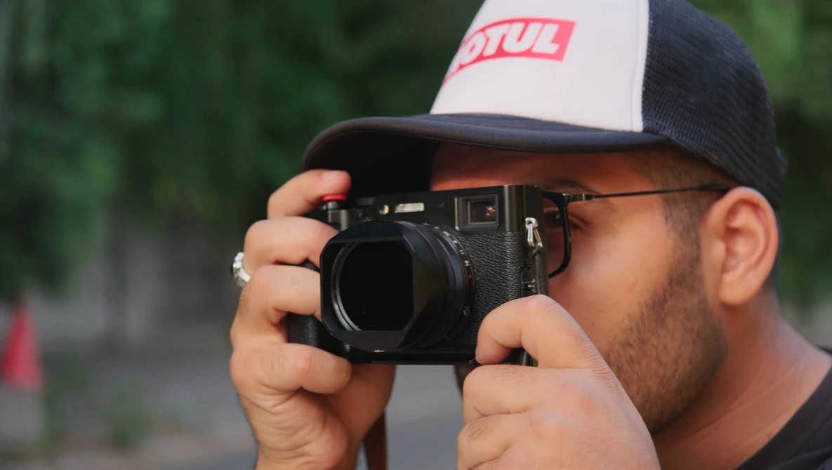 Photographer holding a rangefinder camera up to his face, wearing a trucker-style cap.