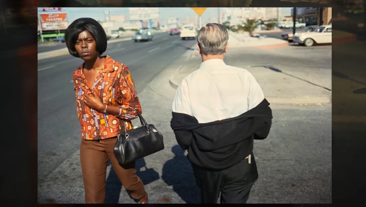 Two people standing on a sunny street with parked cars visible in the background.