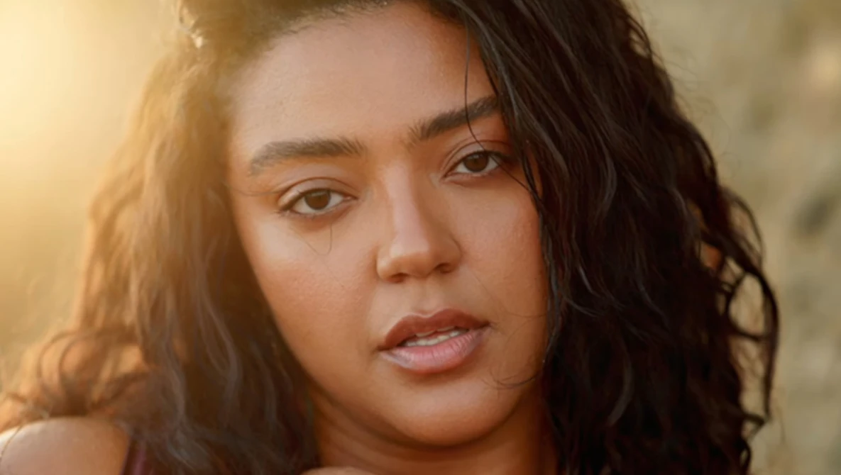 Portrait of a woman with long dark wavy hair, looking directly at camera with warm golden hour lighting.