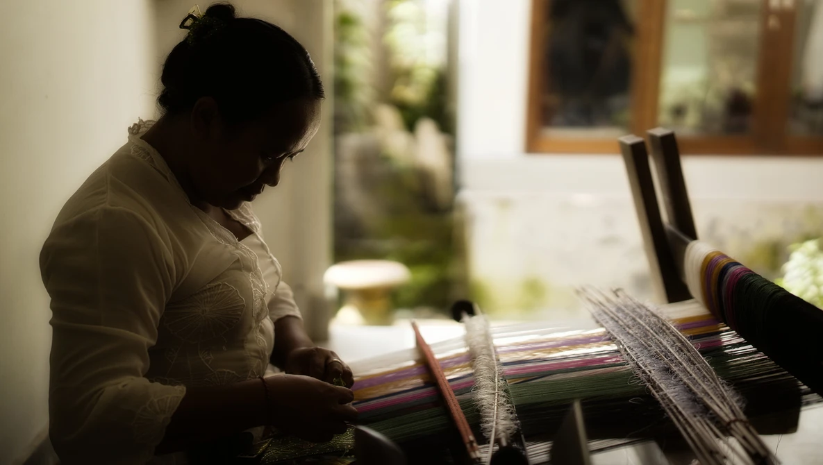 Person working on a weaving loom with colorful yarn threads in soft, directional window light.