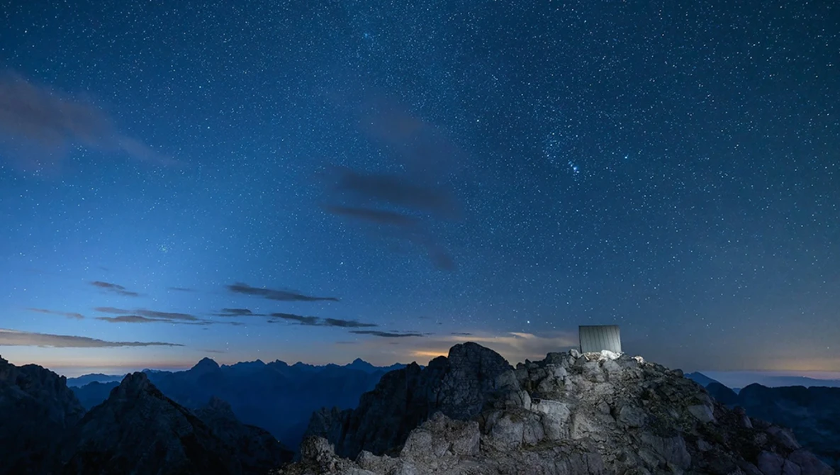 Hilltop castle silhouetted against starry night sky with mountain range in twilight.