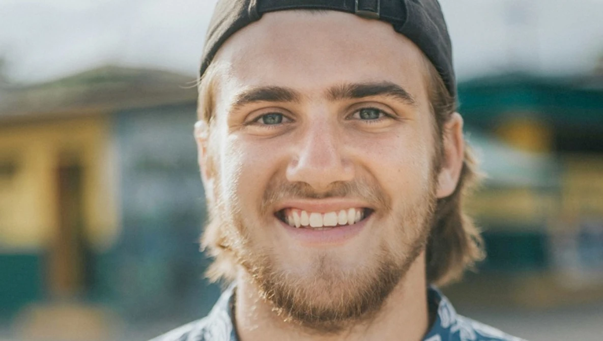 Smiling man with shoulder-length hair and beard wearing a black cap and light colored shirt outdoors.