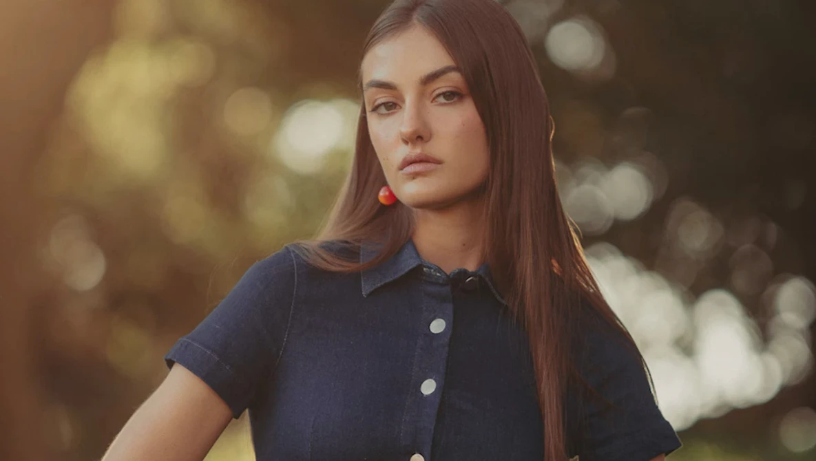 Young woman with long dark hair wearing a navy button-up shirt and red earrings, photographed outdoors with soft golden bokeh background.