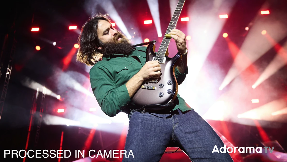 Guitarist performing on stage wearing a green jacket, playing a white electric guitar against red and pink stage lighting.