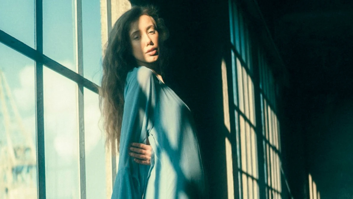 Woman with long dark hair looking over her shoulder by a sunlit window with grid panes.