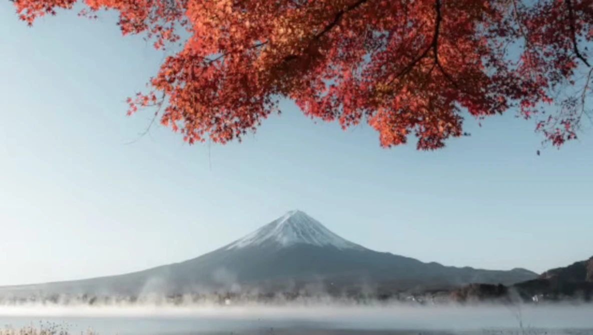 Mount Fuji framed by red autumn maple leaves with morning mist rising from a lake below.