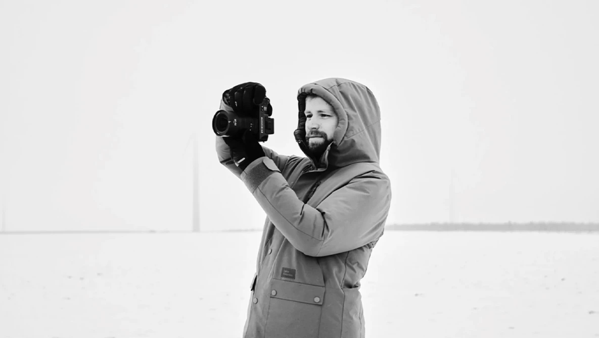 Bearded man in hooded jacket holding vintage film camera to his face against minimal background.