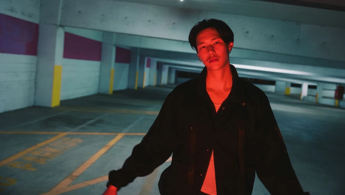 Man in black jacket standing in empty parking garage with red and yellow lighting.