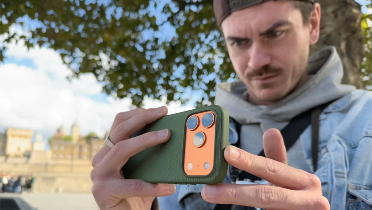 Man holding a green and orange laser rangefinder device outdoors with historic architecture in background.