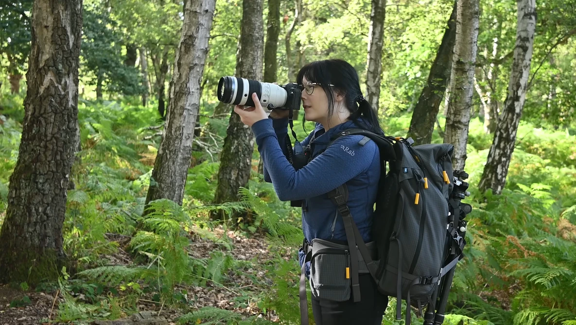 Photographer with telephoto lens and backpack in forest setting.