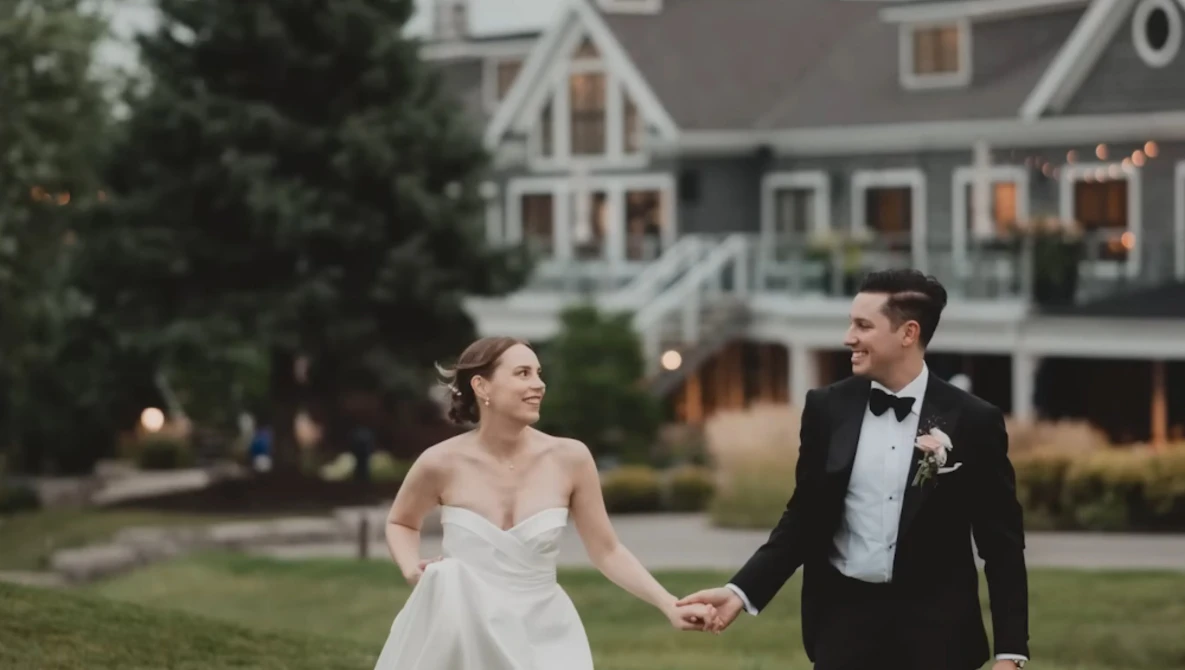 Bride and groom holding hands while walking on manicured lawn in front of large residential home at dusk.