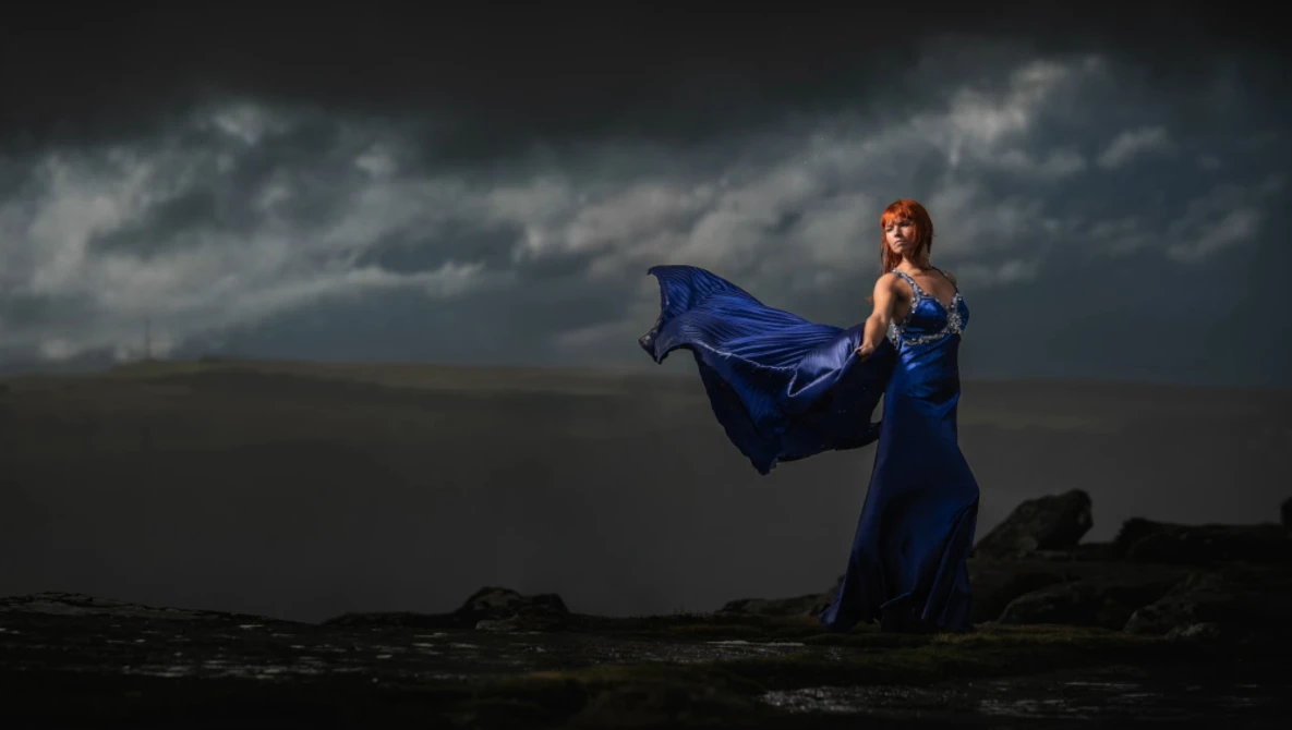 Woman in flowing blue gown standing on rocky shore with dramatic storm clouds overhead.