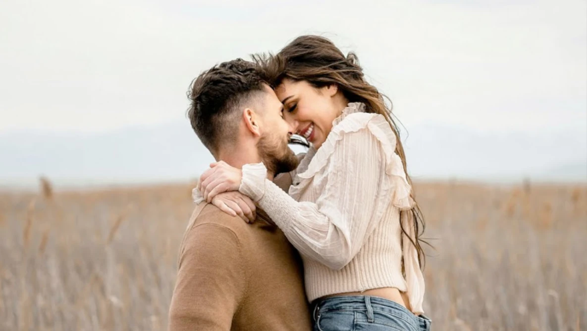 Couple embracing and smiling in an open field with dried grass and overcast sky.