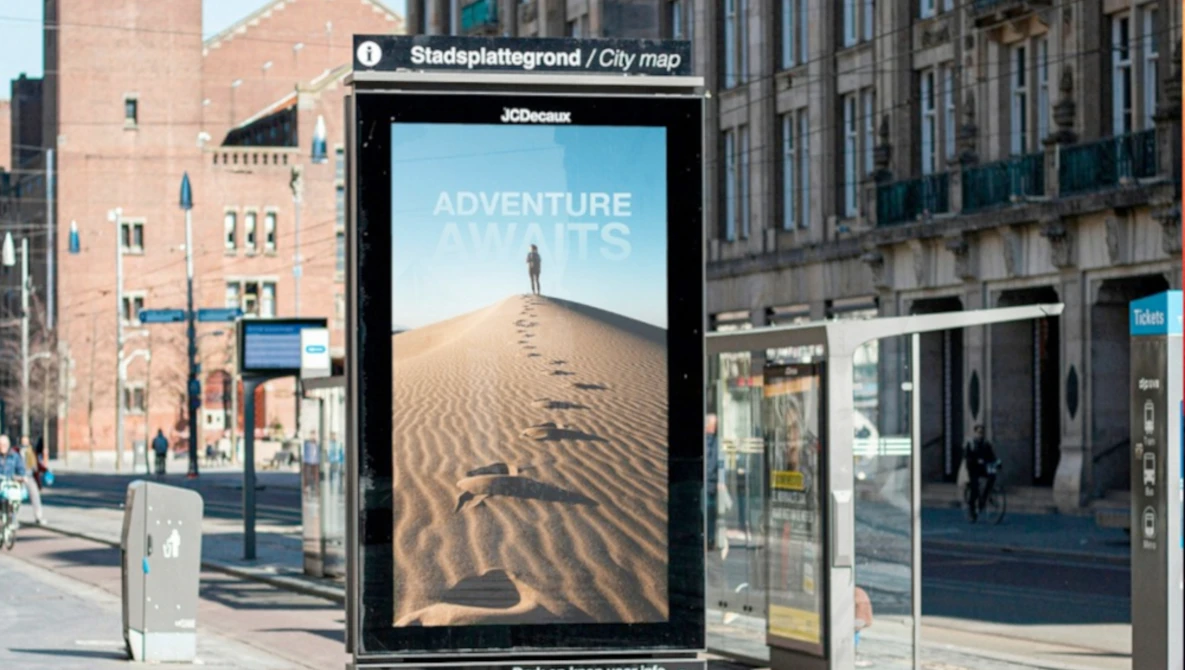 Digital billboard in an urban transit station displaying desert landscape photography with silhouetted figure on sand dune.