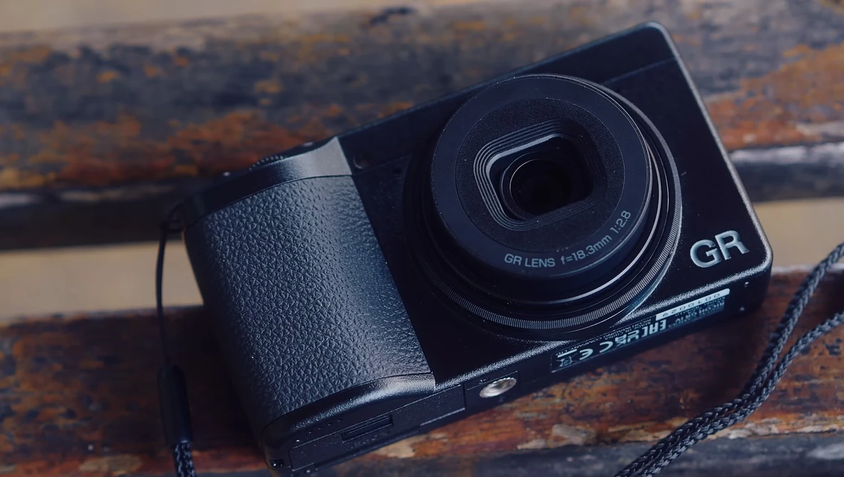 Ricoh GR compact camera with leather grip and neck strap on weathered wooden surface.
