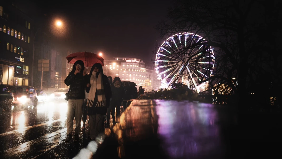 Wet city street at night with illuminated Ferris wheel reflecting in water and pedestrians in foreground.