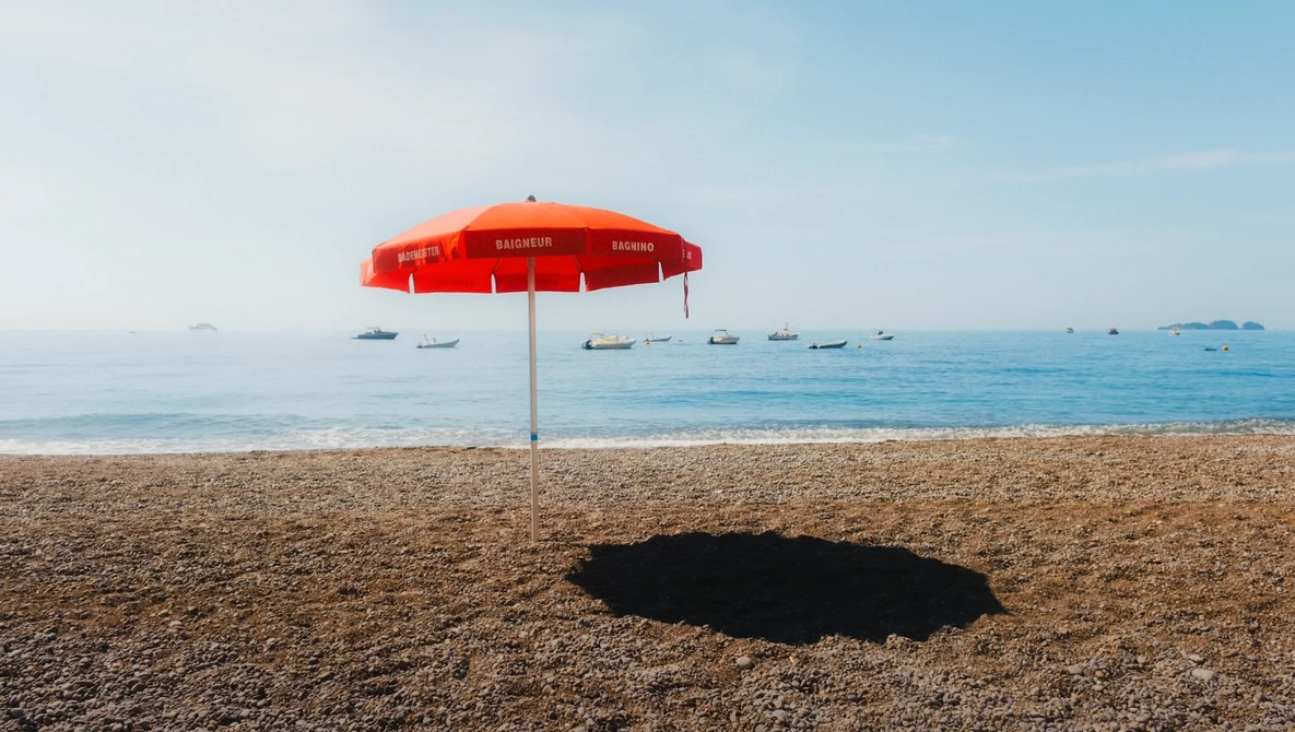 Red beach umbrella casting a dark shadow on sand, with calm ocean and moored boats beyond.