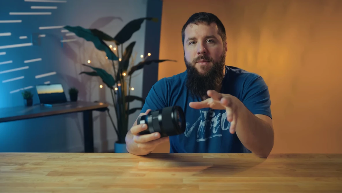 Bearded man in blue shirt demonstrating camera controls at wooden desk with warm backlighting.