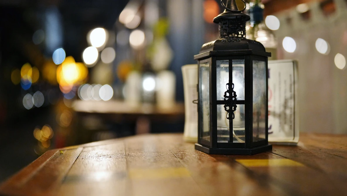 Black metal lantern with glass panels and lit candle on wooden table at night.